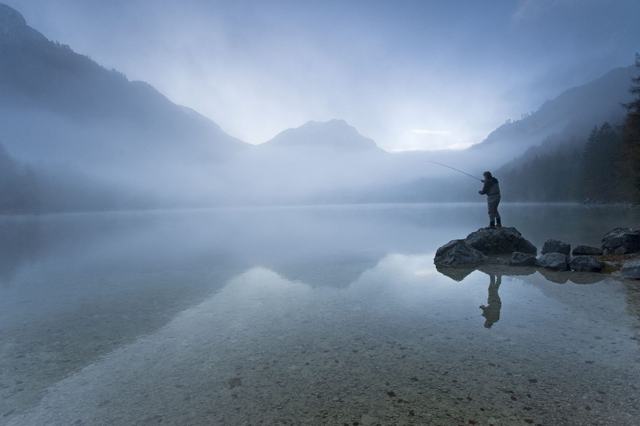 Ein Mensch steht im Morgennebel auf einem Stein im See